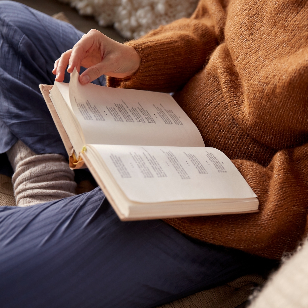 Person in a cozy sweater and blue pants holds a book open while reading.